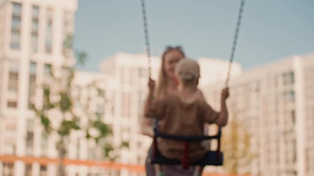 Young boy swings on playground swing while his mother stands nearby watching him. scene is set in urban park with tall buildings visible through soft focus background. perspective emphasizes