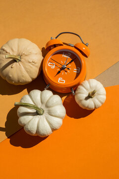 Orange alarm clock with three white decorative pumpkins on two tone beige and orange background for autumn season and daylight saving time concept