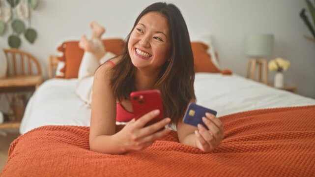 Woman on bed holding smartphone and creditcard, legs raised and smiling while checking screen in a building; online shopping happiness.