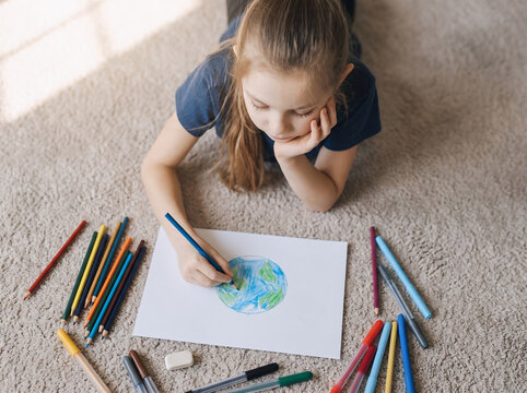 op view of cute child lying on carpet creating artwork of globe with blue and green colors. Concept of children's creativity, environmental awareness, and education.