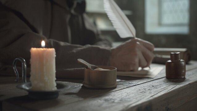 Wooden pounce pot on a rustic table near a lit candle and a monk writing with a quill pen in background inside a scriptorium