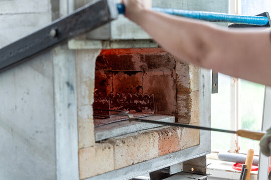 Artisan opening glowing hot muffle kiln oven with metal tool in enamel and ceramic art studio workshop.