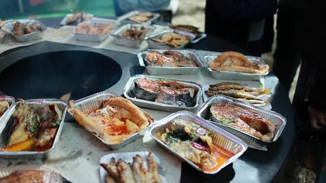 Fresh fish dishes in foil containers on street food table. Grilled salmon steaks and fried small fish with herbs and spices. Seafood street market concept, top view