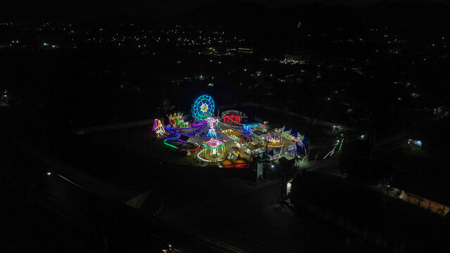 Aerial view of a vibrant night carnival featuring a glowing Ferris wheel and colorful amusement rides illuminated against the dark landscape in Mataram, West Nusa Tenggara, Indonesia.