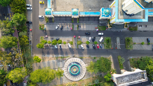 Aerial view of a mosque complex with green domes, a central fountain, and a parking lot filled with cars in Indonesia.