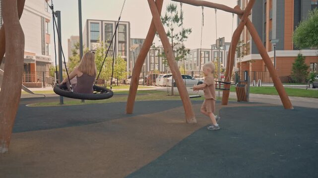 Woman swings on tire swing at outdoor playground while young boy pushes same child friendly equipment. scene takes place in modern urban park with residential buildings green trees