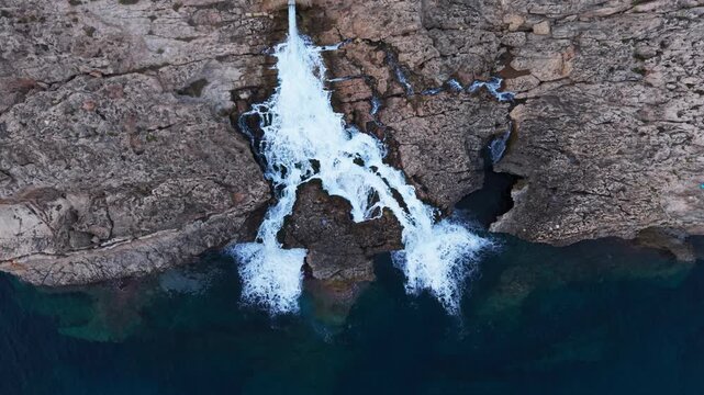 Industrial wastewater cascades from a reverse osmosis desalination plant outlet down rugged limestone cliffs into the sea, captured by drone above Ghar Lapsi, Malta.