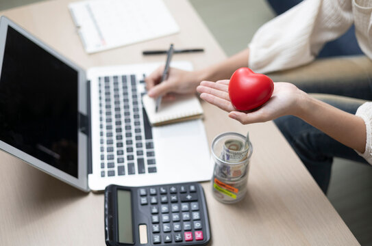 The concept of saving for health and medical expenses is evident as a young woman jots down notes on her savings, holding a red heart symbol in her hand, inside a bright, modern building.