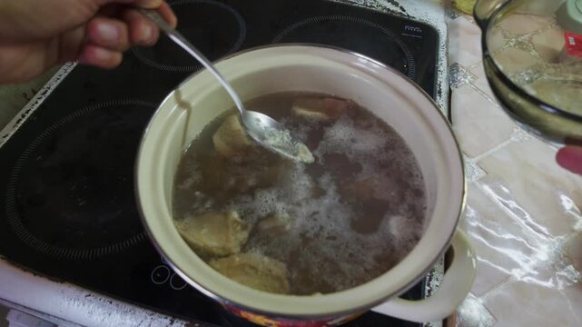 A hand uses a spoon to skim foam from a simmering pot of meat broth on an electric stove. The gentle bubbling liquid is cleaned of impurities while a glass lid rests nearby on the countertop.
