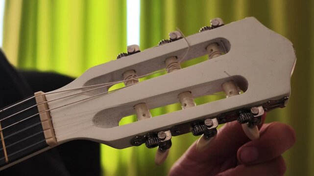 A hand methodically adjusts the tuning pegs on a white classical guitar headstock against a green curtain background, finishing with a steady hold to indicate the tuning process is complete.