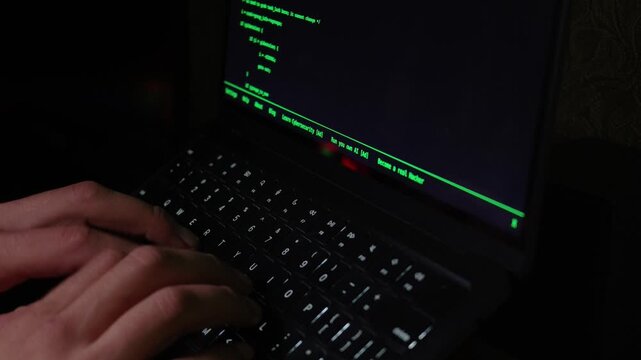 Hands type rapidly on a black laptop keyboard in a dimly lit setting, with green command-line text scrolling on the screen.
