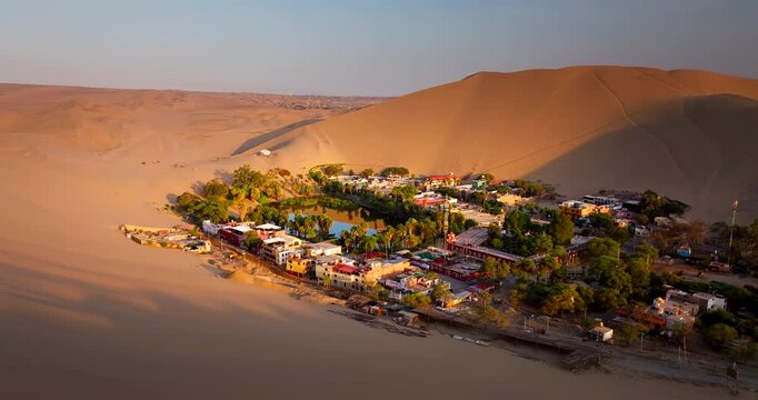 Huacachina oasis and lakeside settlement surrounded by vast desert dunes in Peru at sunrise, drone orbit as light cuts town in half