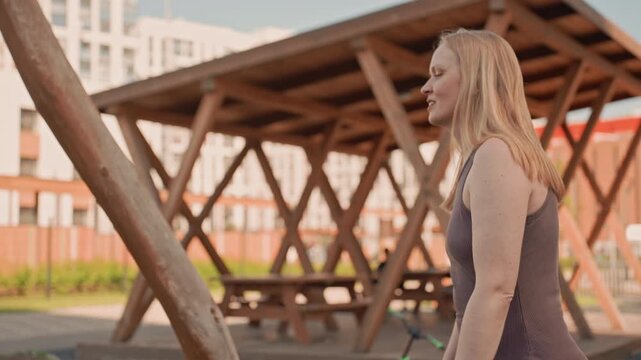 Woman interacts with child on playground equipment gently pushing swing in outdoor park setting. scene features soft natural light and visible background elements like wooden structures