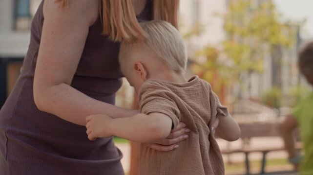 Young boy with light hair interacts closely while being supported by woman in outdoor playground setting. child holds onto play equipment as adult provides physical support emphasizing care