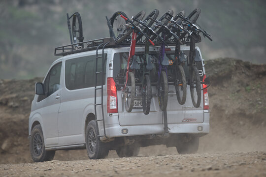 A pickup truck rolls down a dirt trail through lush green hills, its bed packed with five downhill mountain bikes lined up wheel to wheel, race plates and knobby tires facing the camera. 