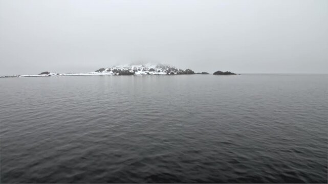 Remote Snowy Island Sits on Calm Gray Svalbard Arctic Sea in Summer. A low rocky island with lingering snow lies on the horizon across smooth gray water beneath a soft overcast polar sky.