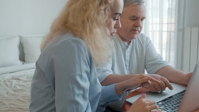 Adult daughter helps her senior father work with laptop and understand online services at home. Utility bill payment, household finances, digital payments, online accounts, technology education