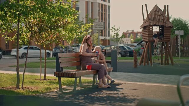 Woman and young boy sit on bench in urban park interacting with each other. scene includes trees parked cars buildings playground equipment like wooden treehouse visible behind them