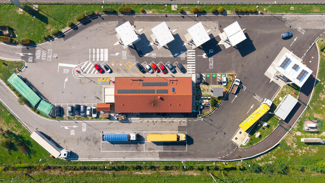 Aerial view of a highway rest stop featuring fuel pumps, parked cars, trucks, and a main building with solar panels, surrounded by green fields in San Giuliano Milanese, Lombardy, Italy.