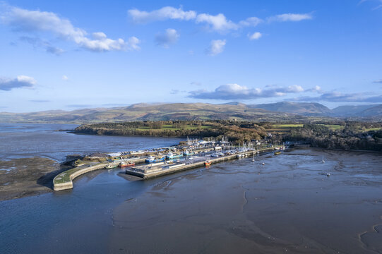 Wales - 27 February 2022: Aerial view of Bangor marina at low tide with exposed mudflats, docked boats, and rolling hills in the background under a clear blue sky.