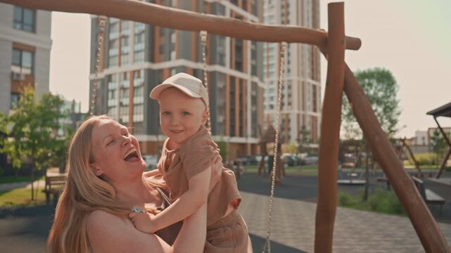 Woman and young boy enjoy time together at playground with mother pushing her child on swing. scene features outdoor park elements like swings and benches against urban backdrop of tall