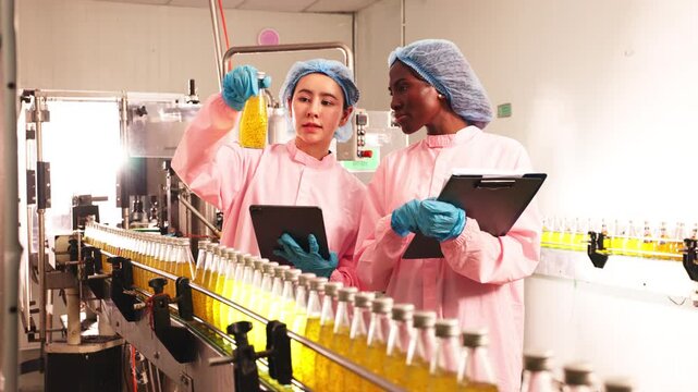 Food factory workers in protective clothing inspecting bottled beverages on production line, recording data for quality control and safety compliance