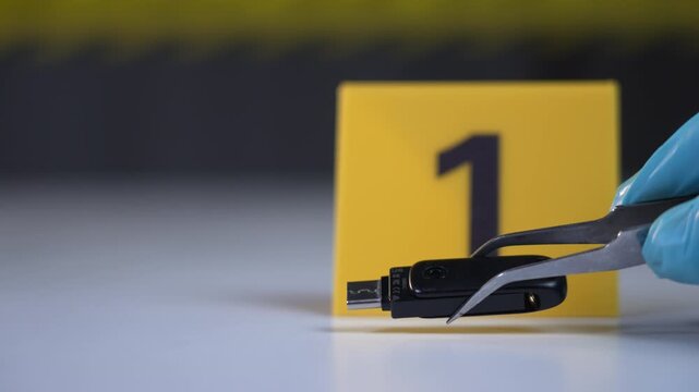 USB drive held by gloved hand using tweezers at yellow evidence marker 1 on white marble surface with police line tape in dark background for cyber crime investigation, side view close up shot