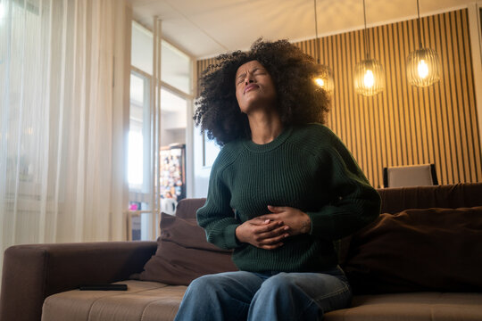 Stress, stomach pain and woman on a sofa with menstruation, gas or constipation, pms or nausea at home. Gut health, anxiety and lady with tummy ache in living room from ibs, bloated or endometriosis
