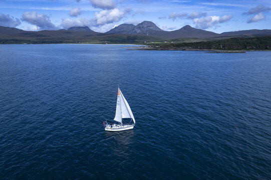 Isle of Jura, Scotland, United Kingdom - 24 August 2022: Aerial view of a sailboat on deep blue waters near the Isle of Jura coast with the Paps of Jura mountains in the background.