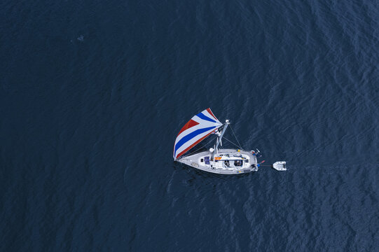 Scotland - 17 August 2022: Aerial view of a sailboat with a colorful spinnaker sailing on deep blue water with a small dinghy in tow.