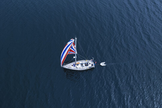 Scotland - 17 August 2022: Aerial view of a sailboat with a colorful spinnaker sail sailing on dark blue water, towing a small white dinghy.