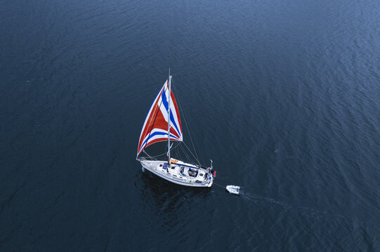 Scotland - 17 August 2022: Aerial view of a sailboat with a colorful Union Jack-patterned spinnaker sail sailing on dark water, towing a small white dinghy behind it.