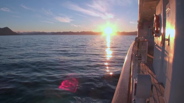 Low Arctic Sun Glows Beside Ship Rail in Svalbard Summer Waters. Golden light reflects across dark water beside the vessel, creating a bright path toward the horizon under broken polar cloud.