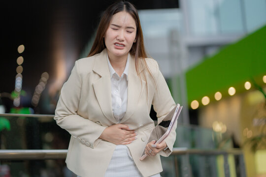 Stomach pain concept of a young businesswoman holding her abdomen with discomfort while carrying a tablet at work. stomach pain, abdominal pain, woman, discomfort, workplace