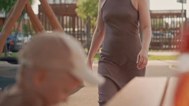 Woman walks through outdoor park setting passing child who is focused on something in front of them. scene features soft daylight and blurred background elements like trees playground