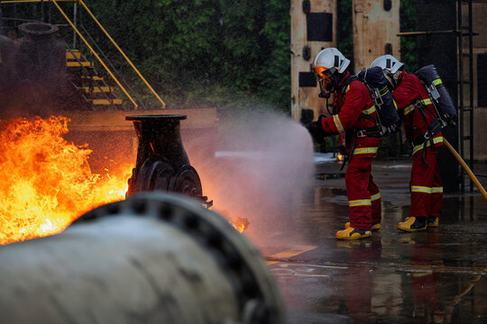 Brave professional firefighters in full protective gear using a high pressure water hose to extinguish the dangerous industrial fire during an intense emergency rescue operation training.