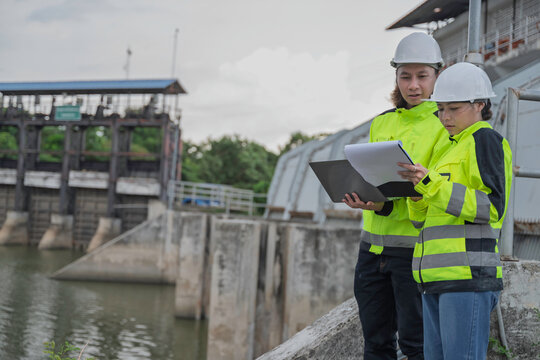Dedicated professional Asian male and female civil engineers collaborate using a laptop and clipboard to inspect heavy water gate dam infrastructure for environmental safety flood control.