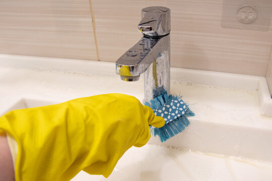 A hand in a yellow glove cleans the sink with a blue brush on the handle. The concept of homework, disinfection
