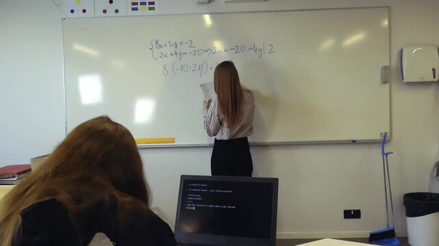 Young female student watching her teacher solve a mathematical equation on a whiteboard in a classroom, with an open laptop showing a programming interface on the desk in the foreground