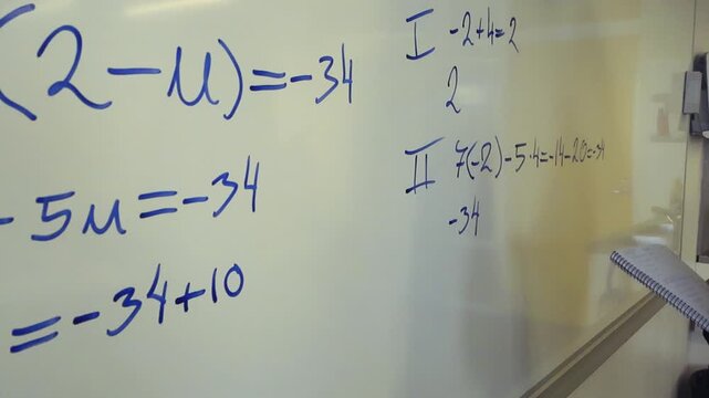 Young female student holding a notebook while writing and solving a complex mathematical equation with a blue marker on a whiteboard during a school lesson, demonstrating the steps of a calculation