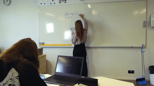 Young female teacher writing an algebraic equation on a whiteboard during a math lesson, while a student watches and uses a laptop with a programming interface on the screen