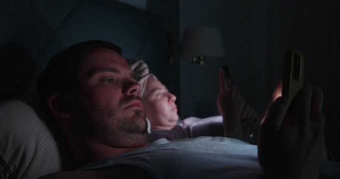 Couple lying in bed and using smartphones before going to sleep at night, representing screen time, social media addiction, insomnia, technology and unhealthy bedtime habits.