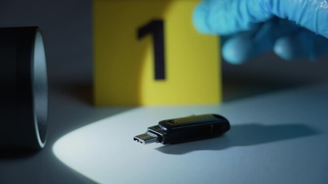 USB flash drive under flashlight beam as hand places evidence marker one, focus shift to marker, dark cybercrime scene, forensic investigation