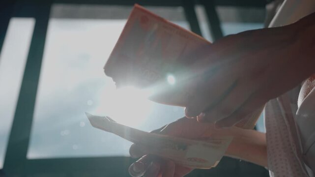 Unrecognizable woman's hands meticulously counting a large sum of 5,000 russian ruble bills, with sunlight creating a bright lens flare against the window background