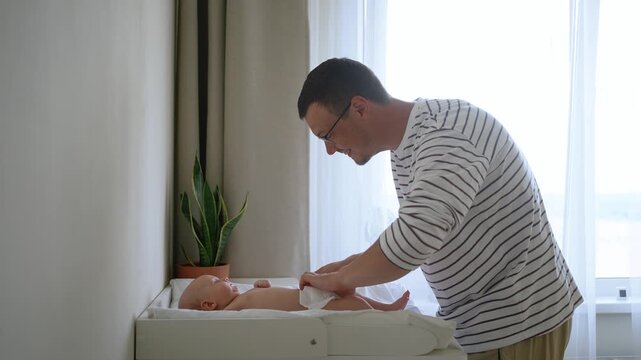 Careful young father putting on a clean new diaper for his adorable infant son lying on a changing table, illustrating modern fatherhood, daily childcare, and family affection