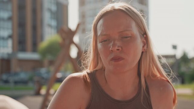 Woman with long blonde hair sits outdoors in park wearing brown tank top. background features modern buildings and greenery under bright daylight. Her expression is neutral as woman looks