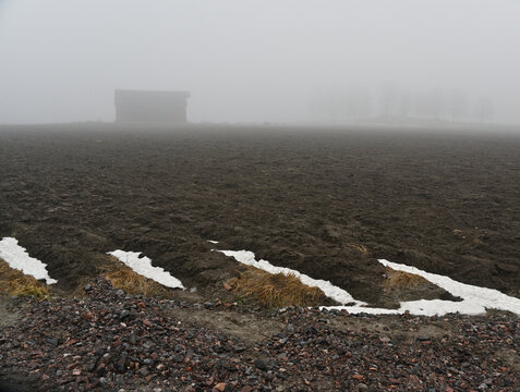 A dark plowed agricultural field covered in dense fog, with patches of melting snow in the foreground and a solitary farm building fading into the mist in the distance
