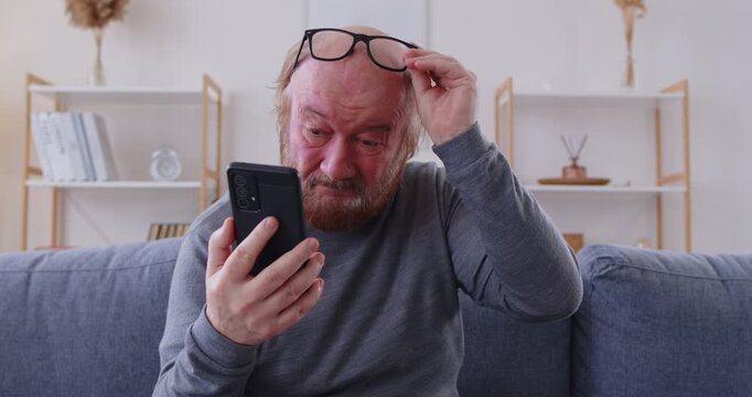 Retired old grandfather with poor vision can't see text on mobile gadget device. Senior man with eyesight problems, sitting on sofa at home, looks at phone, takes off glasses and squints his eyes