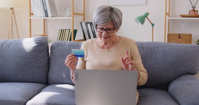 Mature woman using laptop computer and credit card for shopping online, making purchasing in internet, engaged in digital commerce, doing digital transactions, posing on comfortable sofa