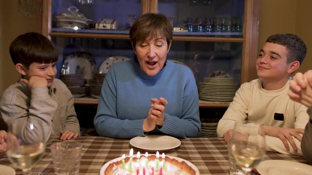 Grandmother closes her eyes to make a wish before blowing out her birthday candles, her two grandchildren watching beside her at the table. A cherished family tradition. Slow motion, video 4K